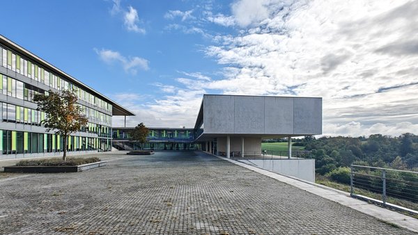 View across the campus courtyard. Several geometric modern building sections made of concrete, glass and coloured surfaces are framed by a green, expansive landscape.